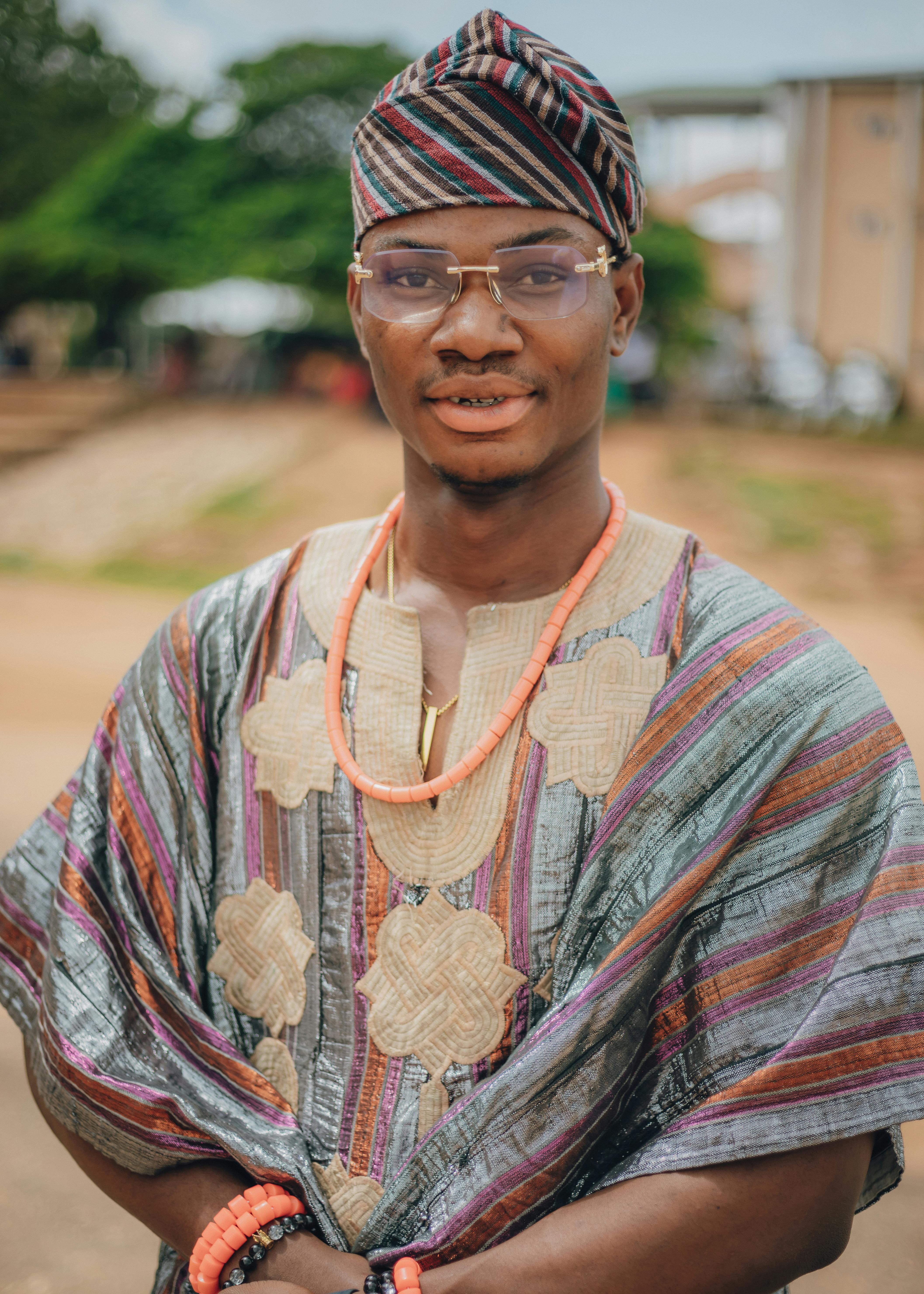 A young man wearing colorful traditional African clothing and beads smiles confidently outdoors.