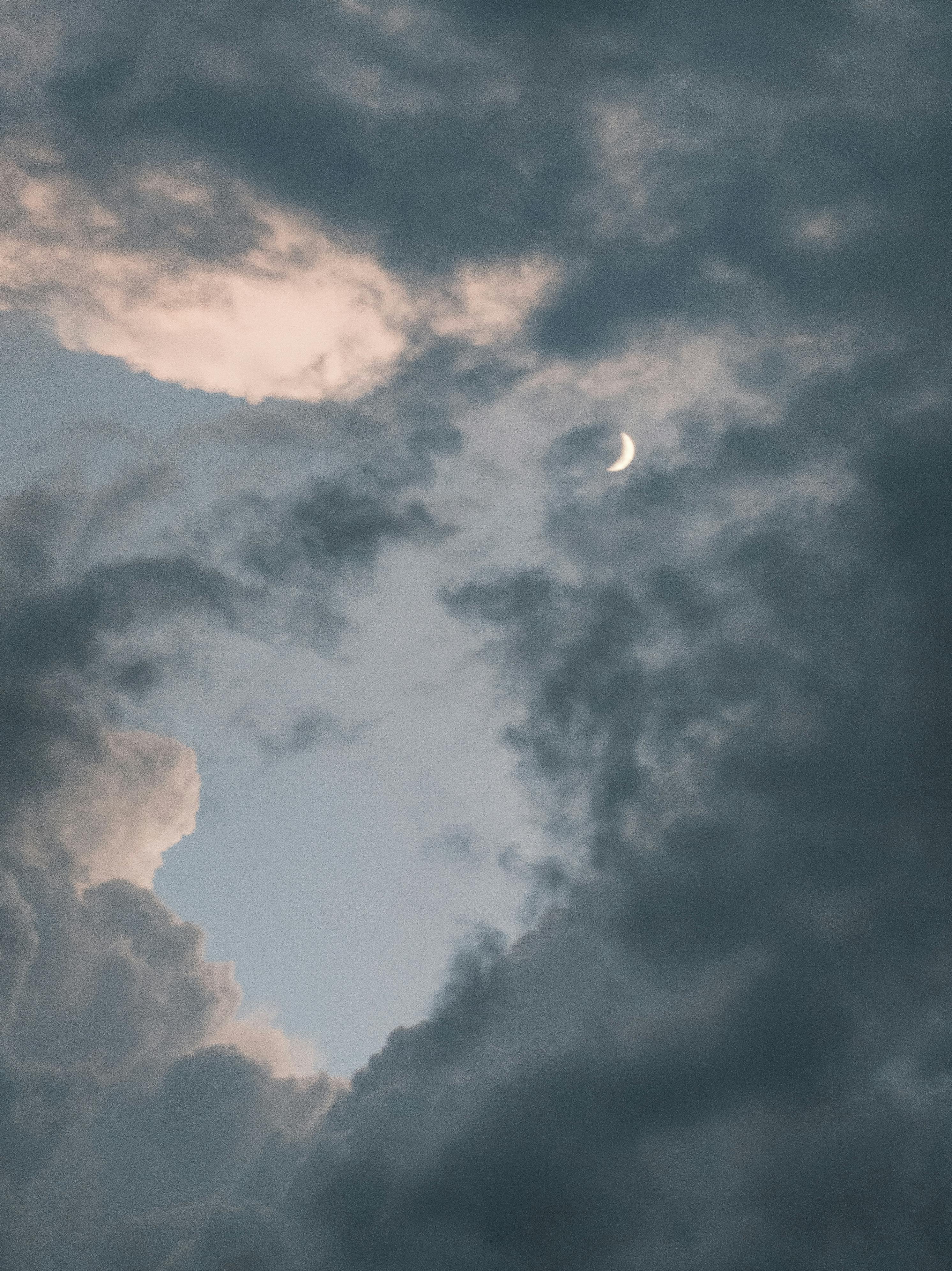 A crescent moon peeks through dramatic, dark clouds during twilight, creating a mystical atmosphere.
