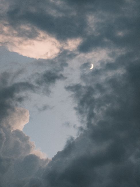 A crescent moon peeks through dramatic, dark clouds during twilight, creating a mystical atmosphere.