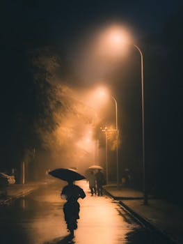 Silhouettes walking with umbrellas on a rainy night illuminated by warm street lamps.