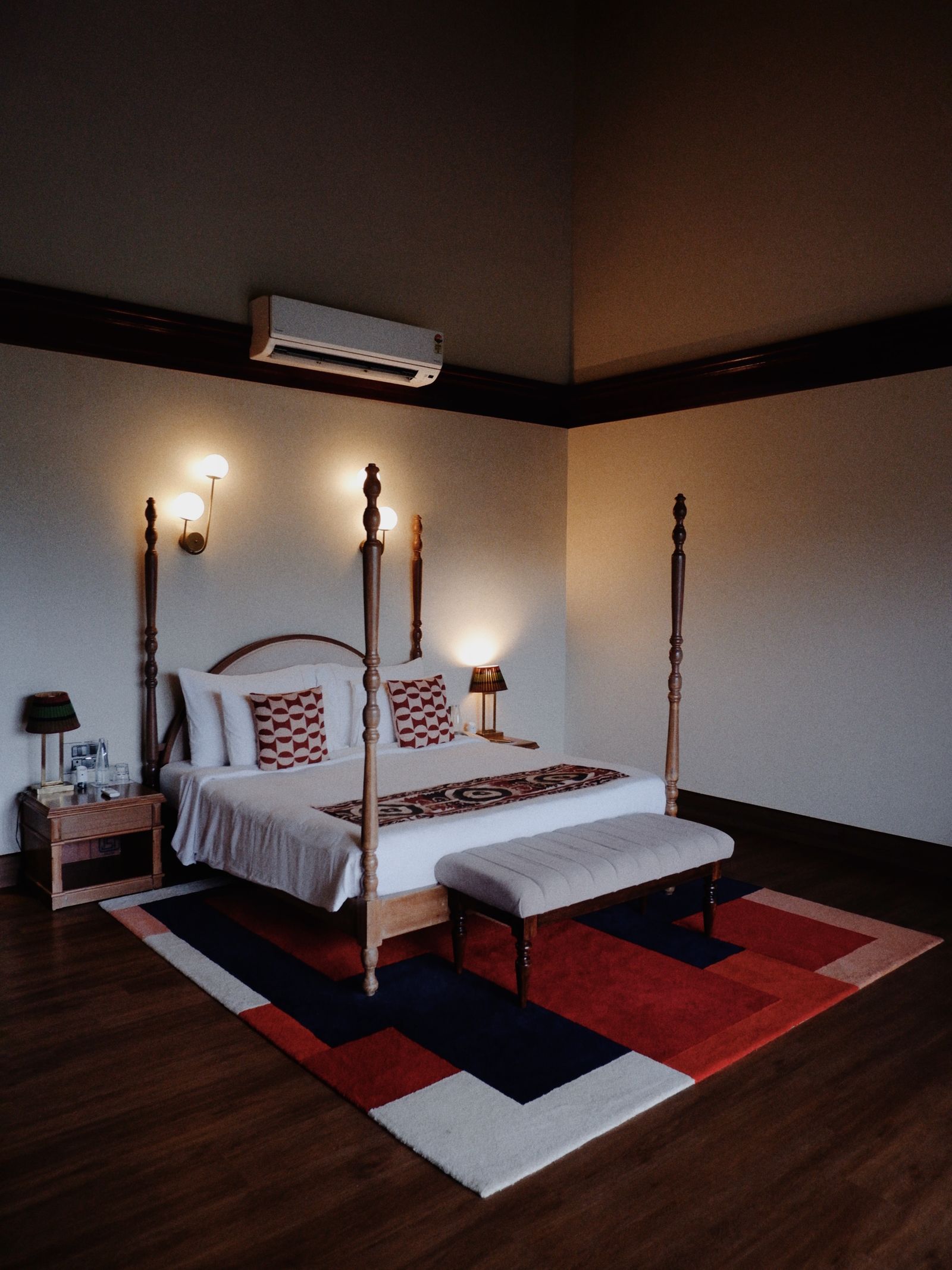 A wooden four poster bed made up in white linen under warm lamplight, a patterned rug on the dark floor