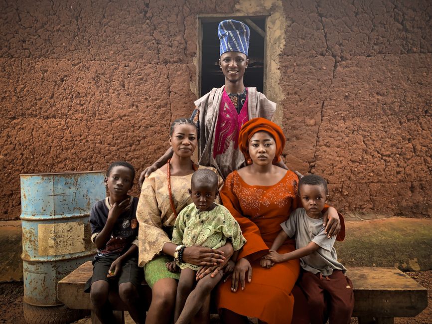 Dark-skinned Nigerian family — mother, father, and two children — neatly arranging large airtight storage drums and food sacks labelled rice, garri, and beans in a tidy kitchen pantry in an Ibadan home, Ankara curtains visible in the background, natural window light, photorealistic