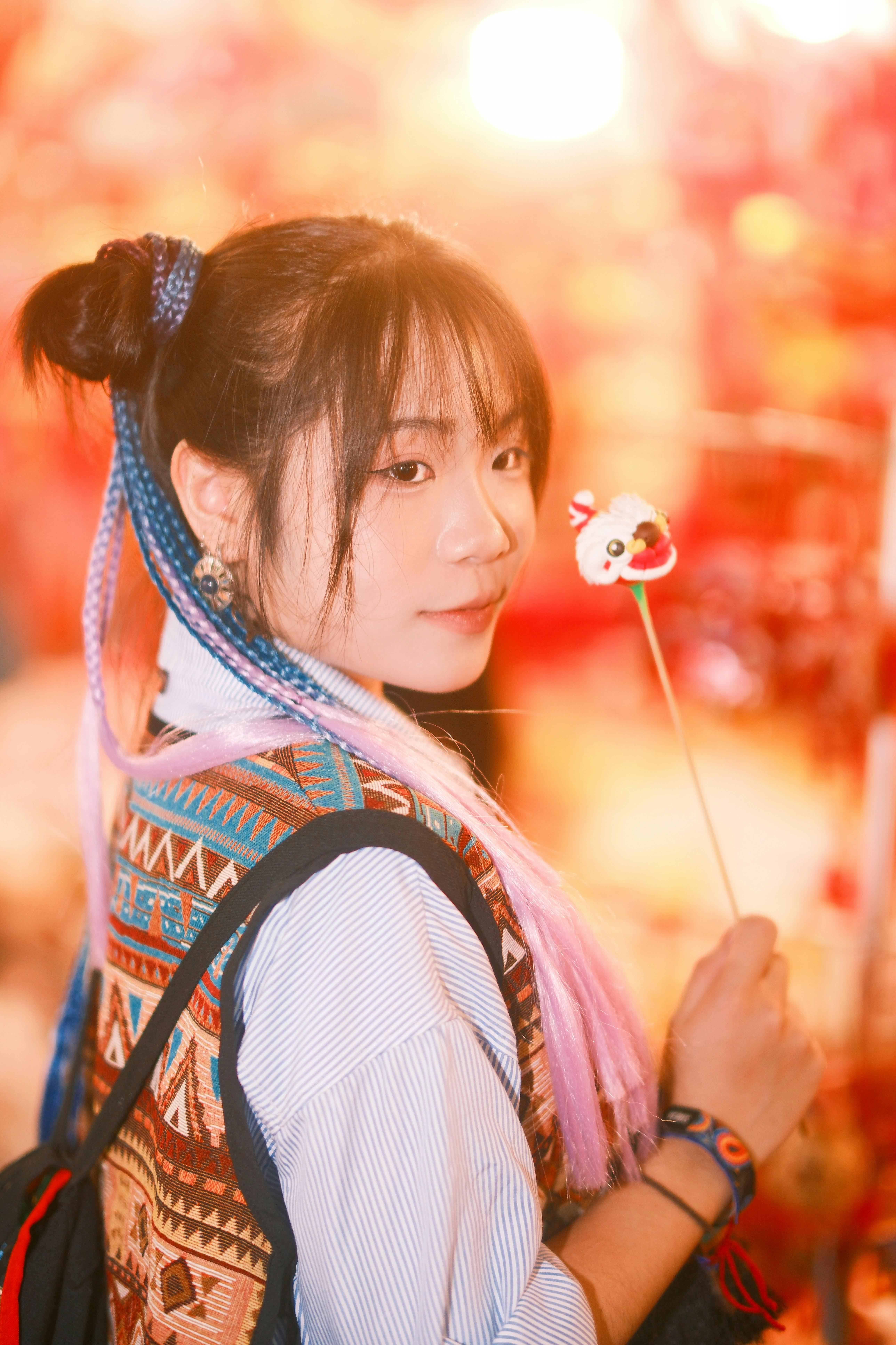 A young woman with colorful hair at a lively festival, holding a stick with a cute decoration.