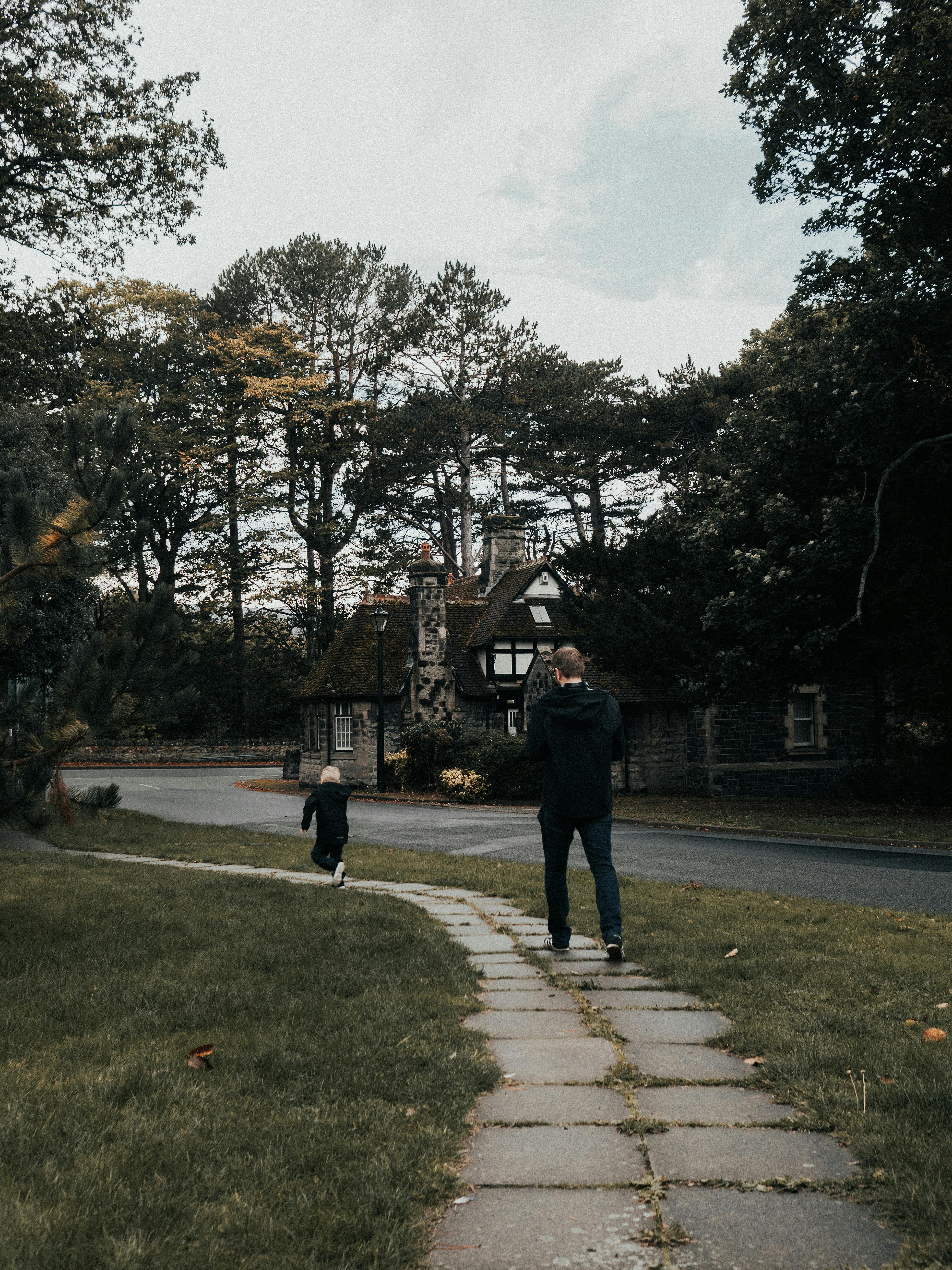 A father and child walk along a path in front of a charming country cottage surrounded by trees.
