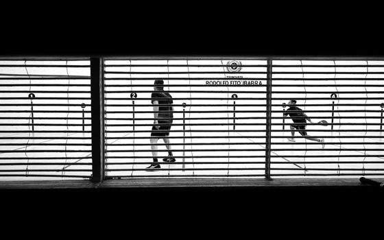 Black and white photo of a squash game through a grate, capturing urban sports activity.