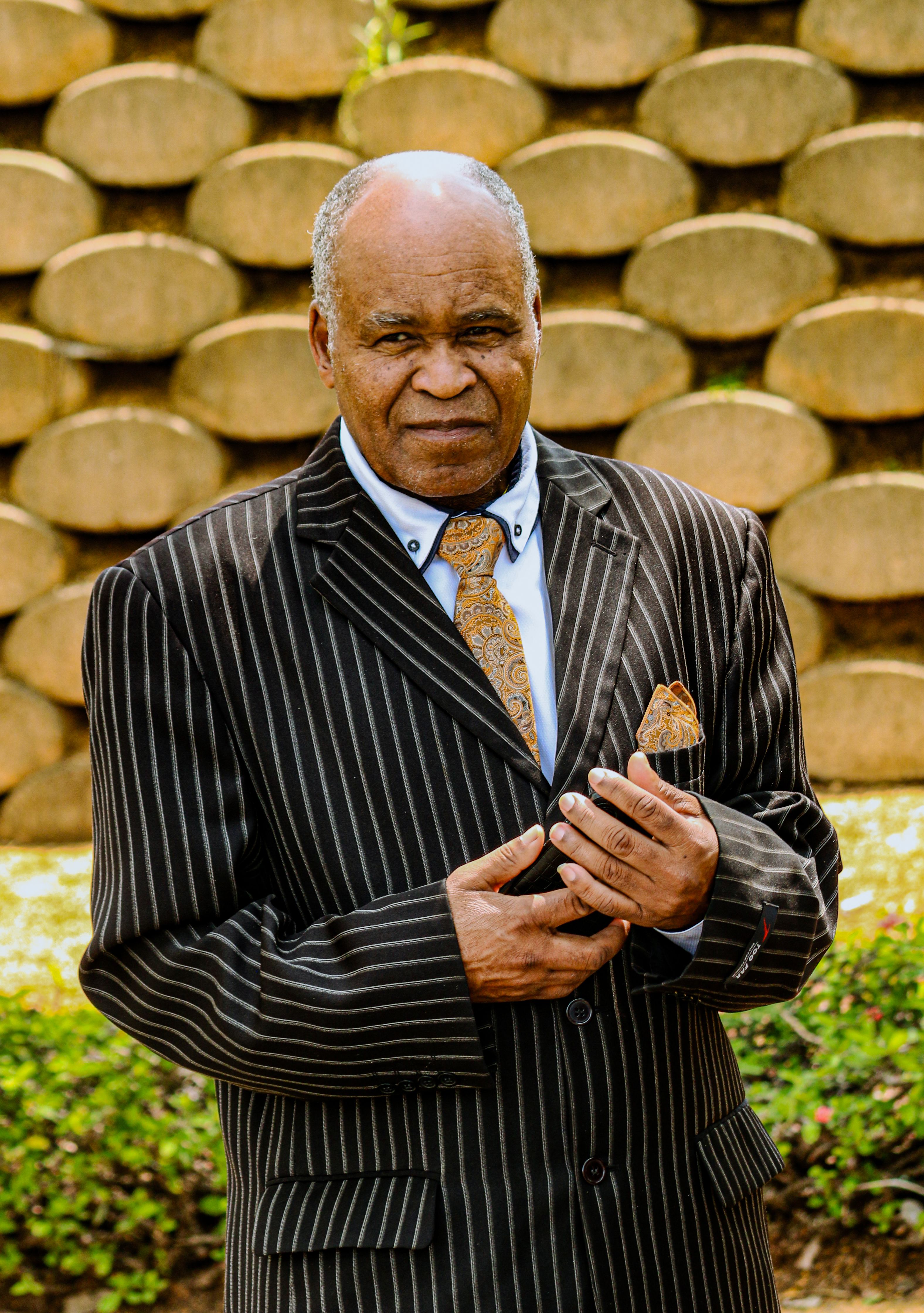 Elderly man in a pinstripe suit poses confidently outdoors against a textured stone wall in Eswatini.