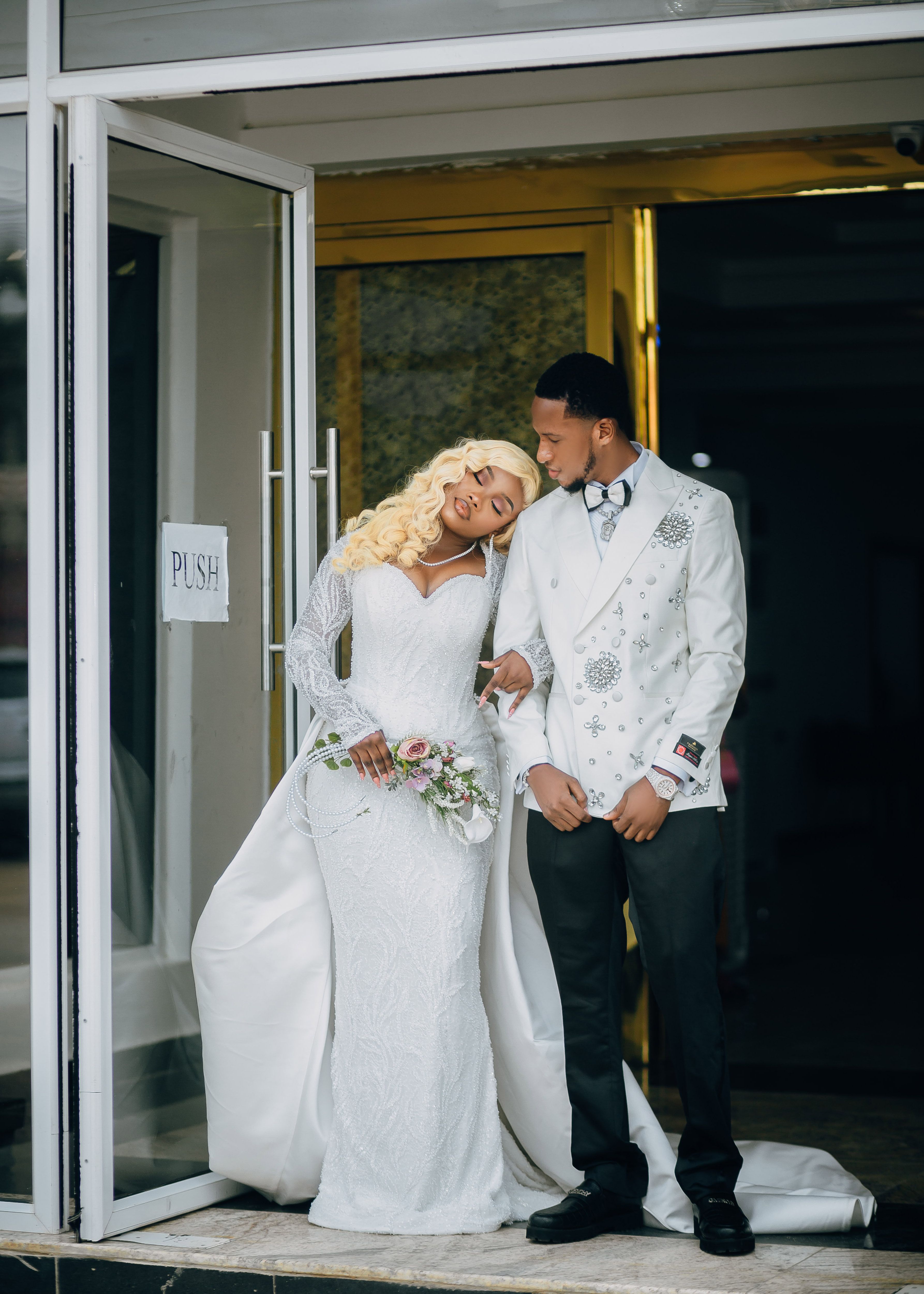 Elegant bridal couple in white attire leaving the wedding venue, sharing a tender moment.