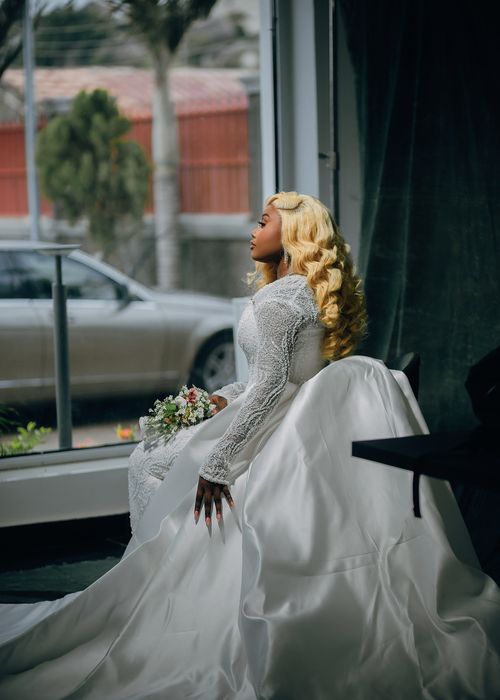Free A beautiful bride in a white gown gazes peacefully out of a window, holding a bouquet. Stock Photo