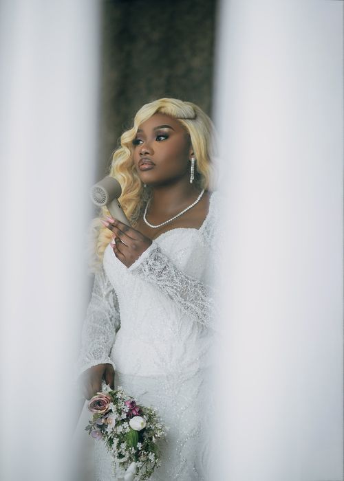 Free Bride in white lace dress holding a bouquet and hair dryer, preparing for wedding. Stock Photo