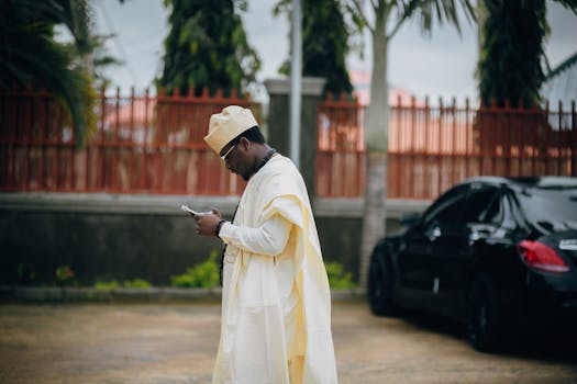 Man in traditional attire using smartphone outdoors beside black car and fence.