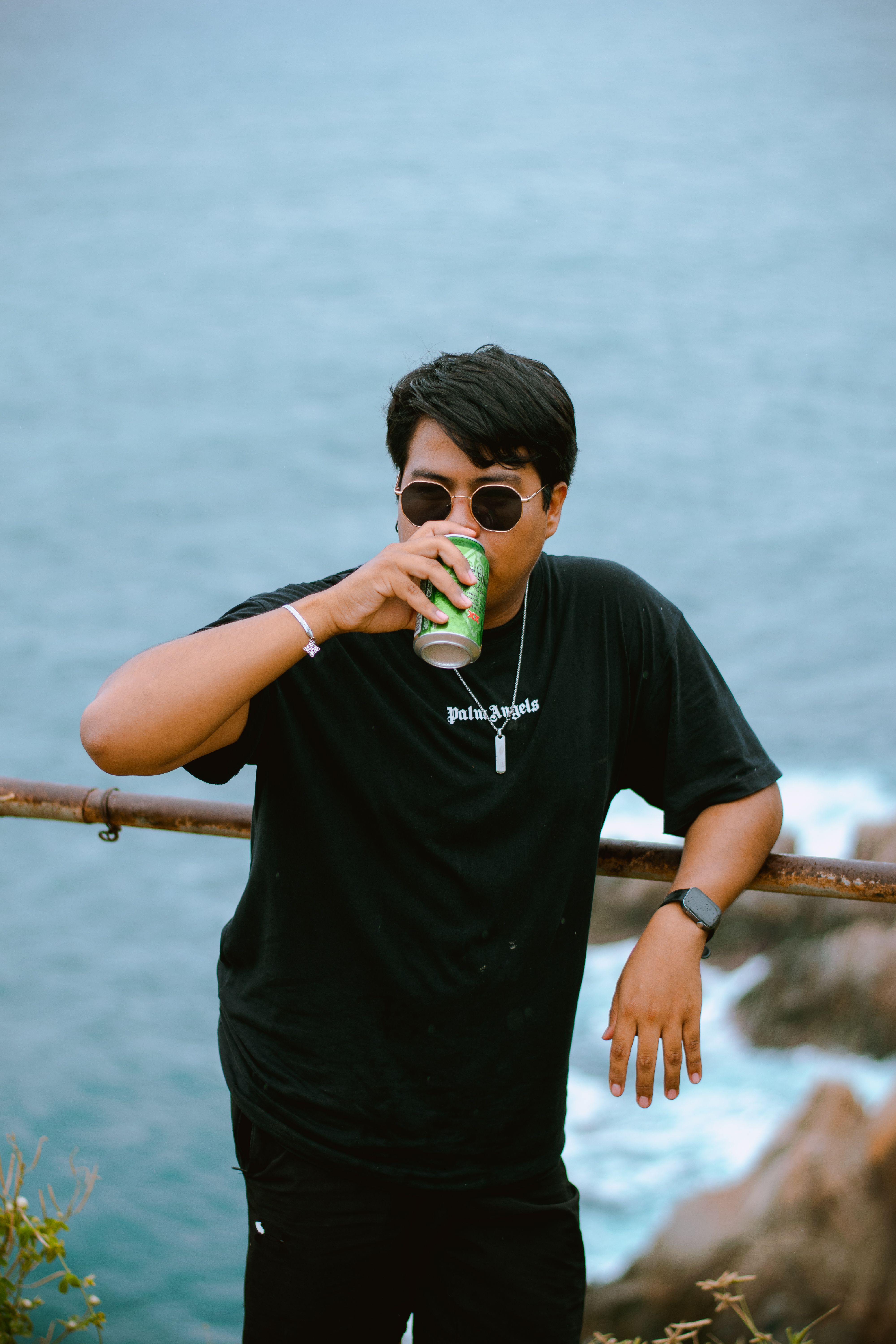 Casual young man wearing sunglasses, drinks from can by the sea.