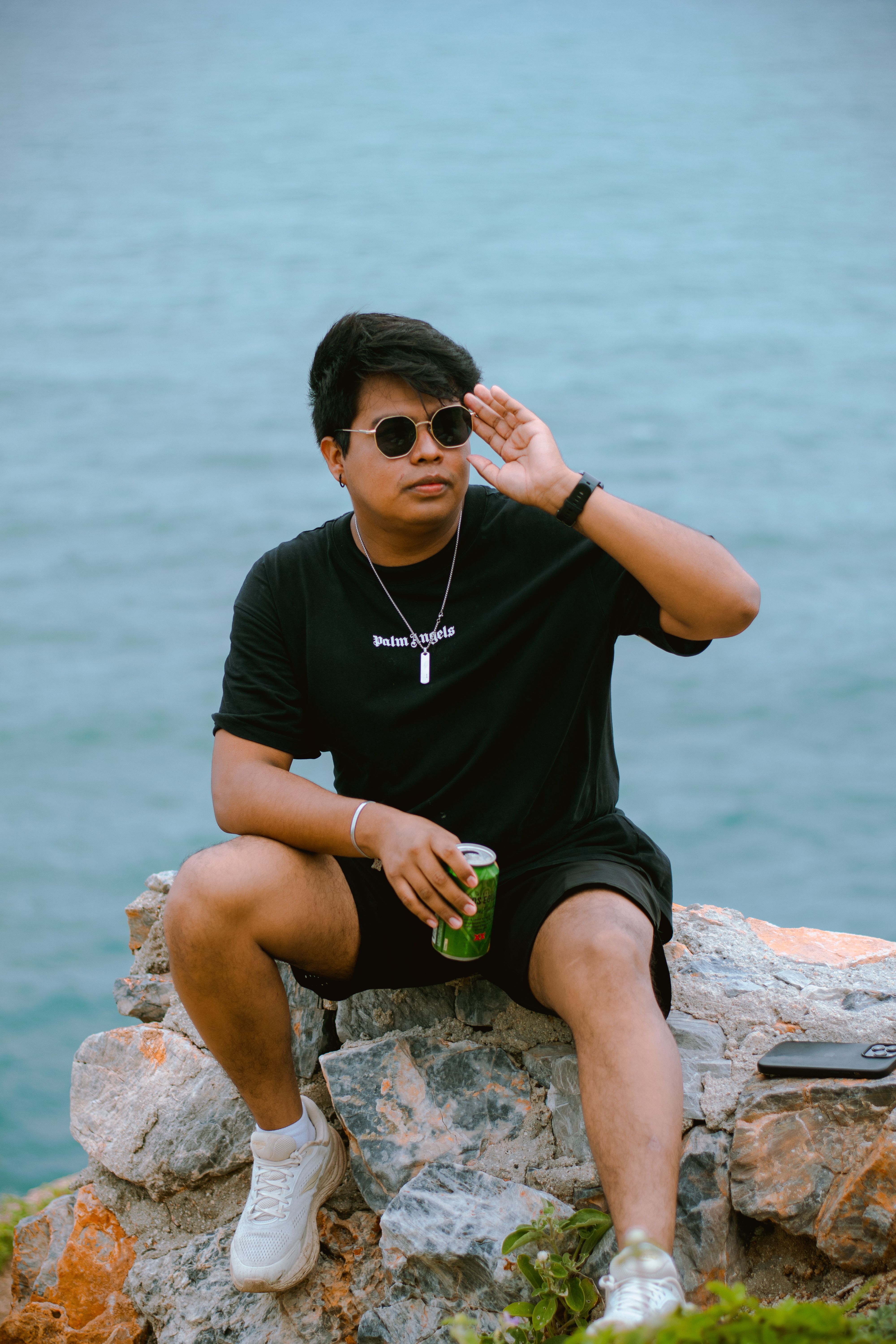 Young man in sunglasses sitting on a rock by the sea, enjoying a drink.