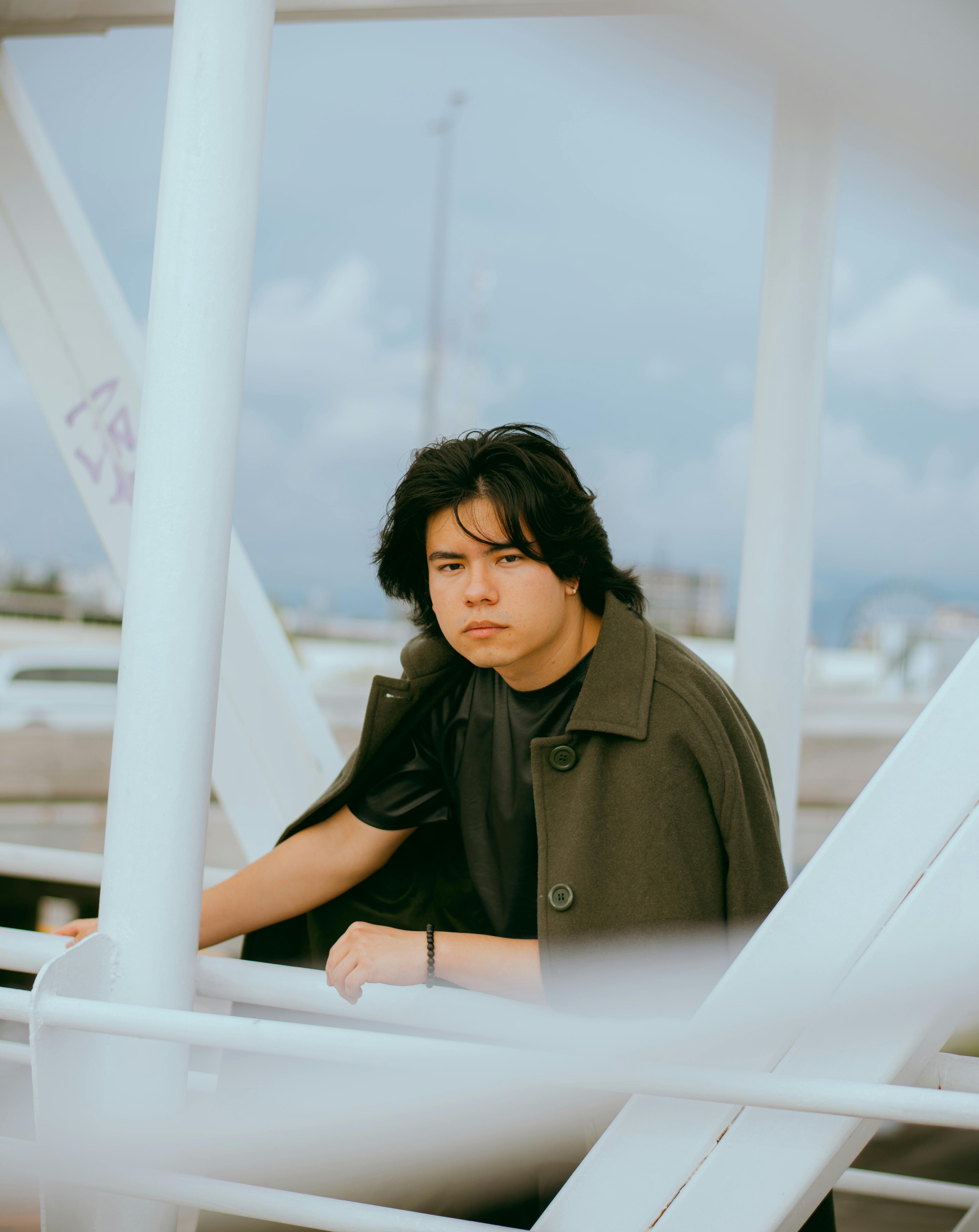 Young man in coat leaning on a bridge railing, reflecting urban vibe.