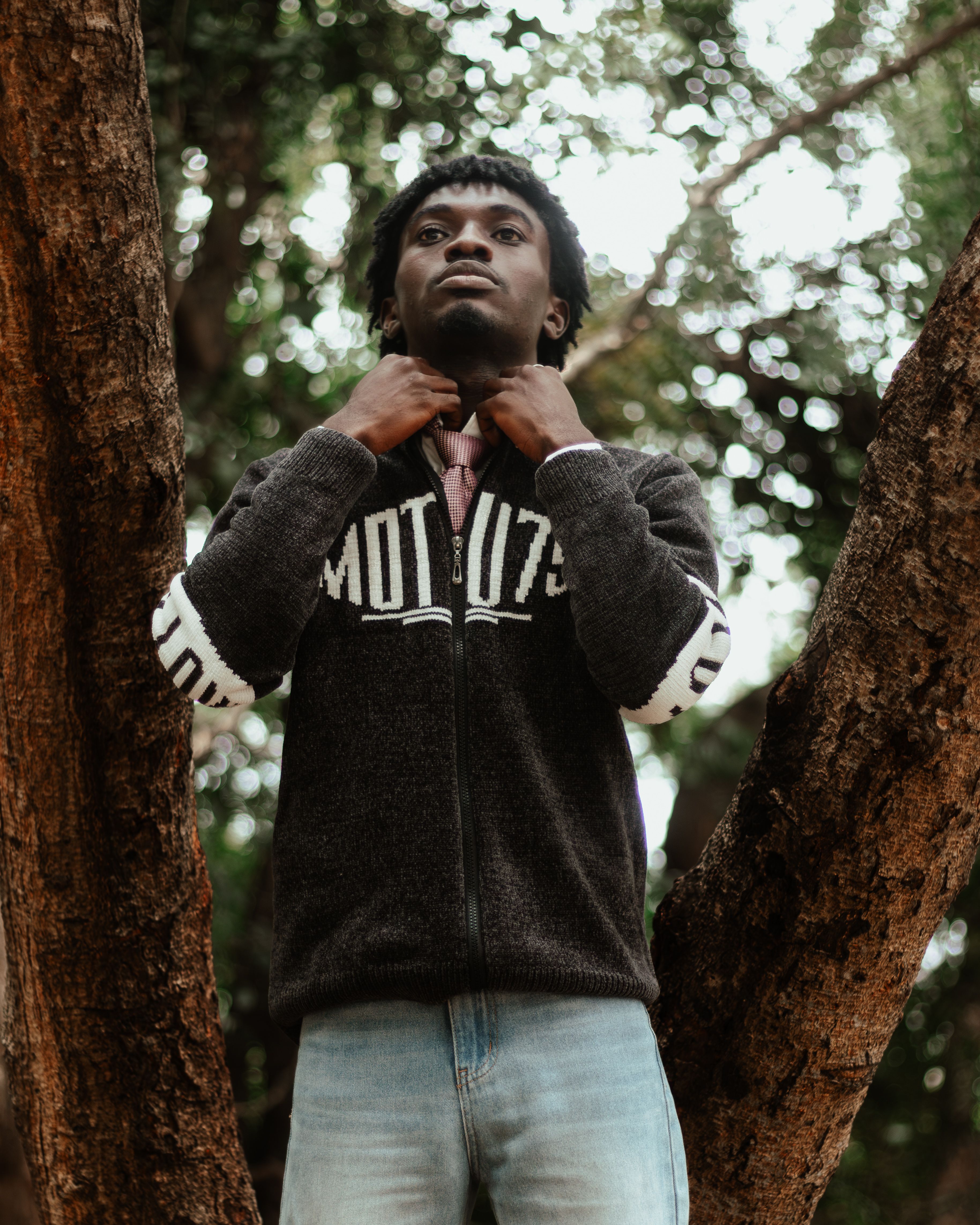 A young man standing between trees, adjusting his collar confidently outdoors.