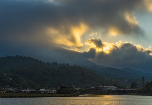 Breathtaking sunset with golden rays piercing dramatic clouds over a lush mountain and lake.