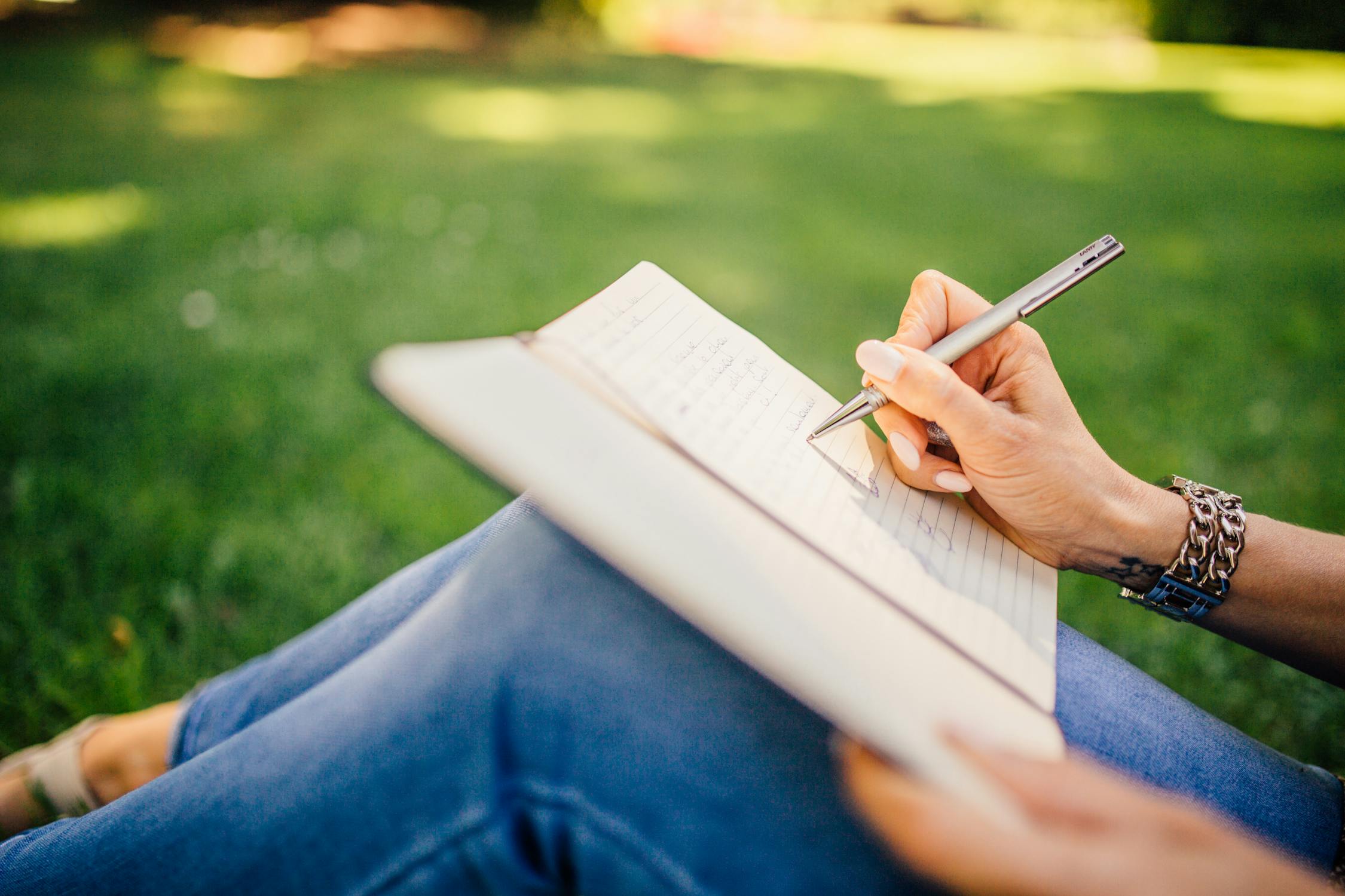 woman writing with pen in her journal while sitting in the grass