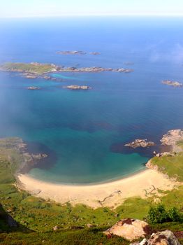 A breathtaking aerial view of a sandy beach and the clear blue ocean, surrounded by rocky coastlines and lush greenery.