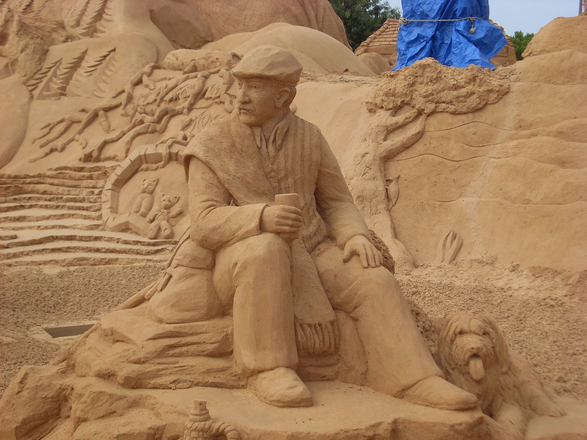 A detailed sand sculpture depicts an elderly man seated with a dog, showcasing artistic craftsmanship.