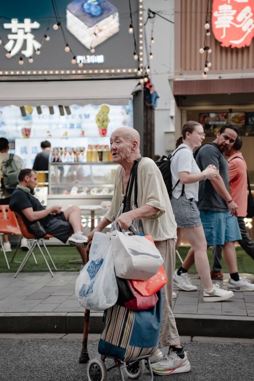 Free Elderly man with bags on a street in an urban shopping area, people walking and conversing. Stock Photo
