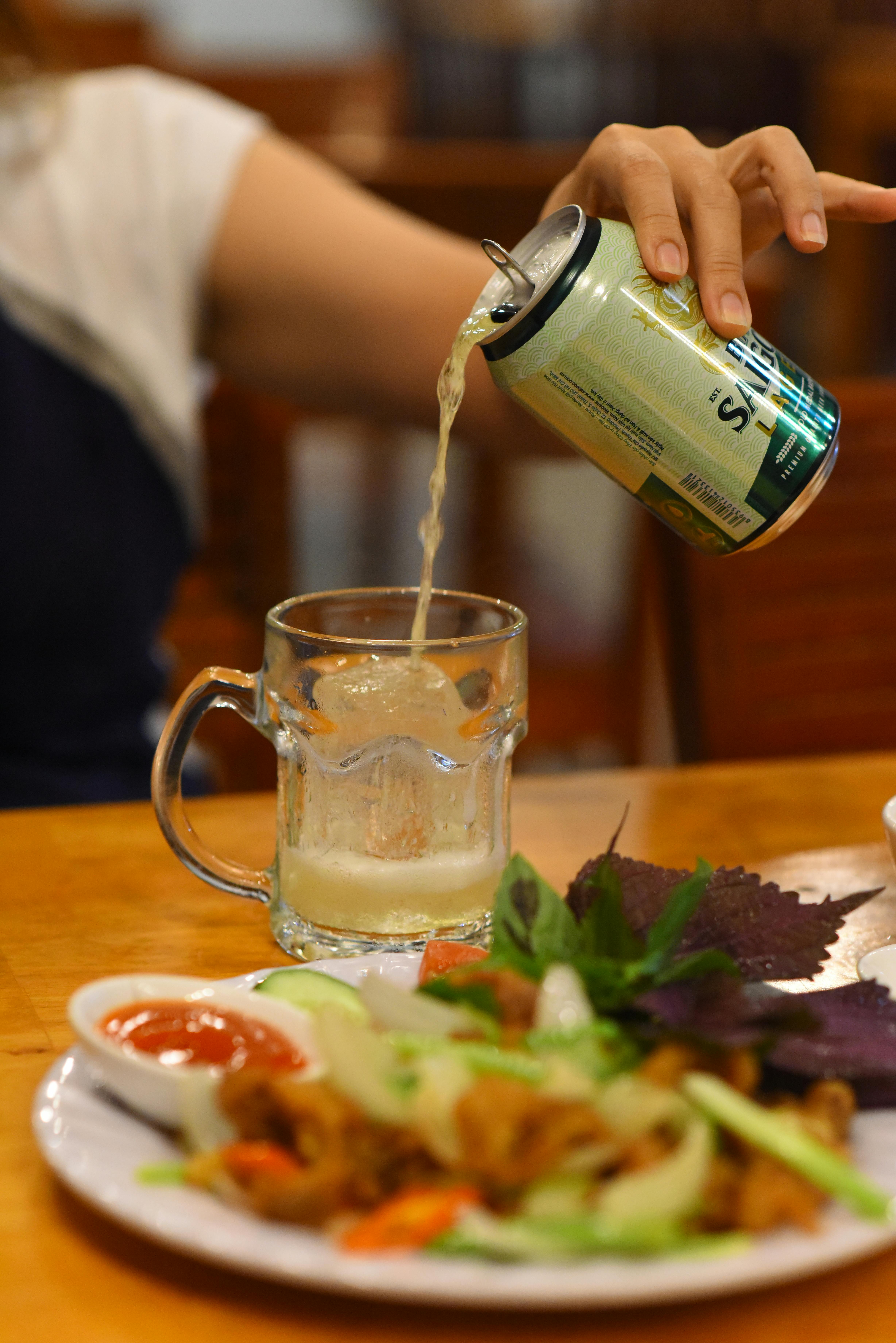 A hand pours a can of lemon-lime soda into a glass beside a plate of salad and sauce.