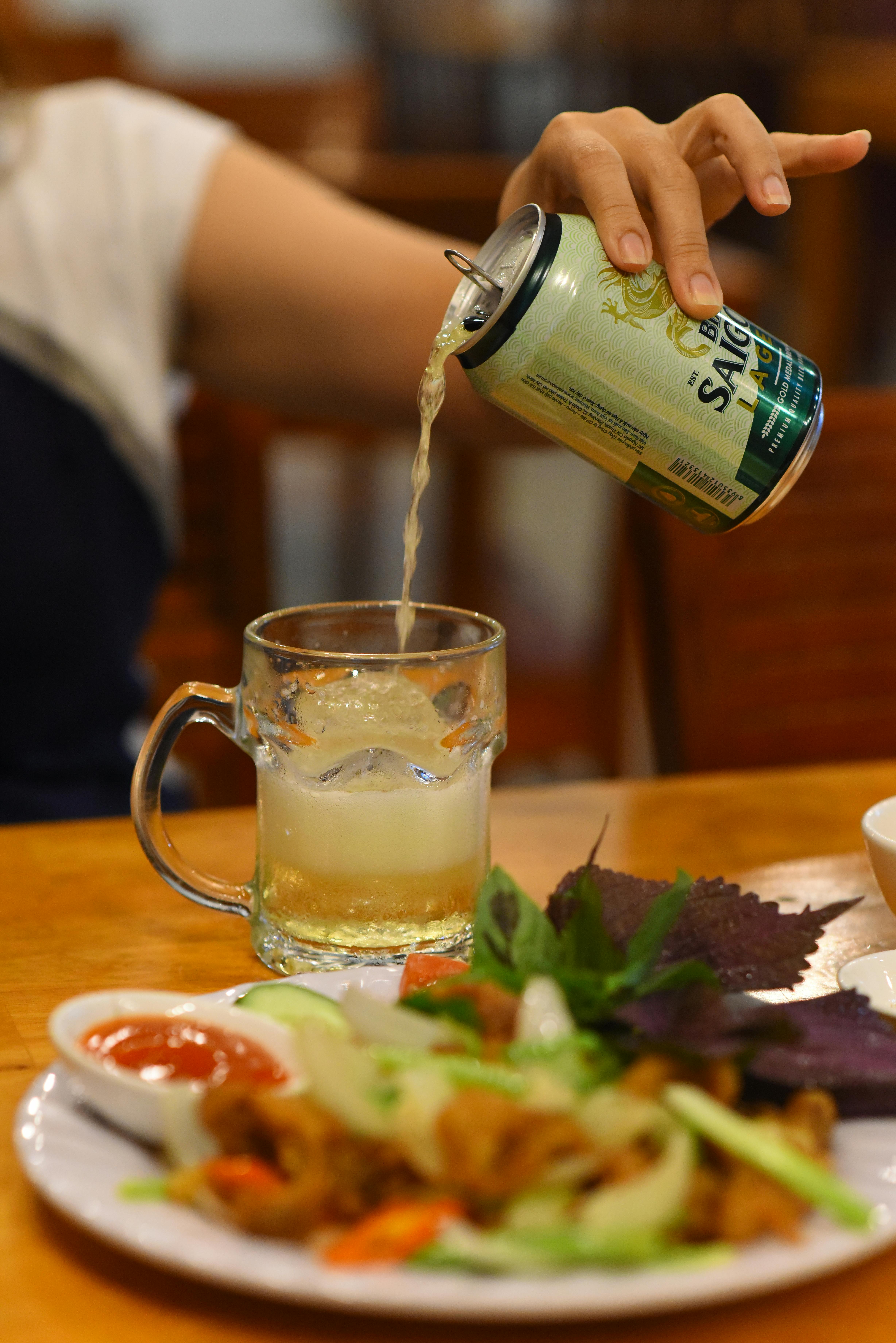 Hand pouring beer into a glass beside a traditional Vietnamese meal with herbs and vegetables.