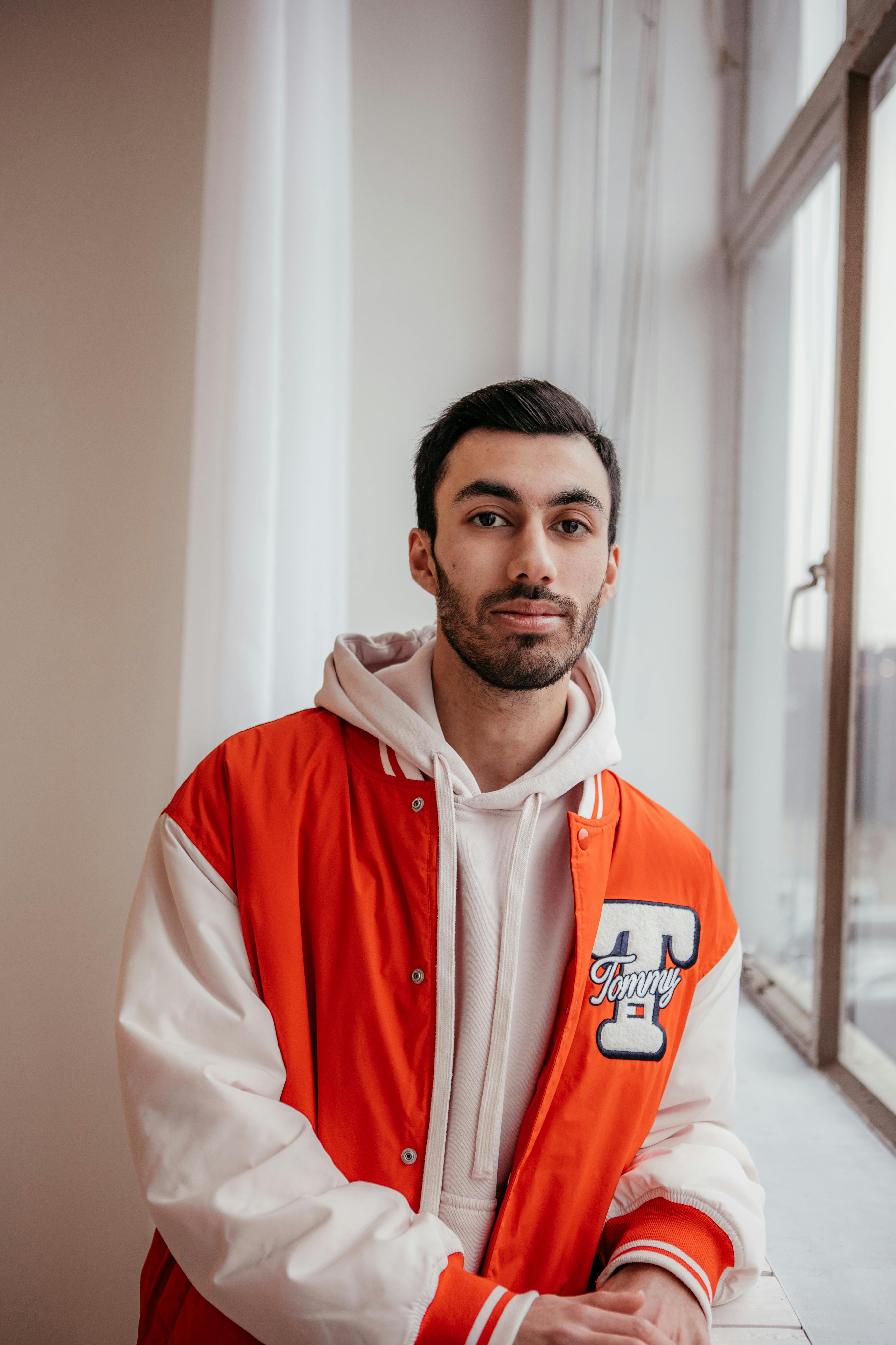 Casually dressed man in a vibrant red jacket poses by a window indoors.