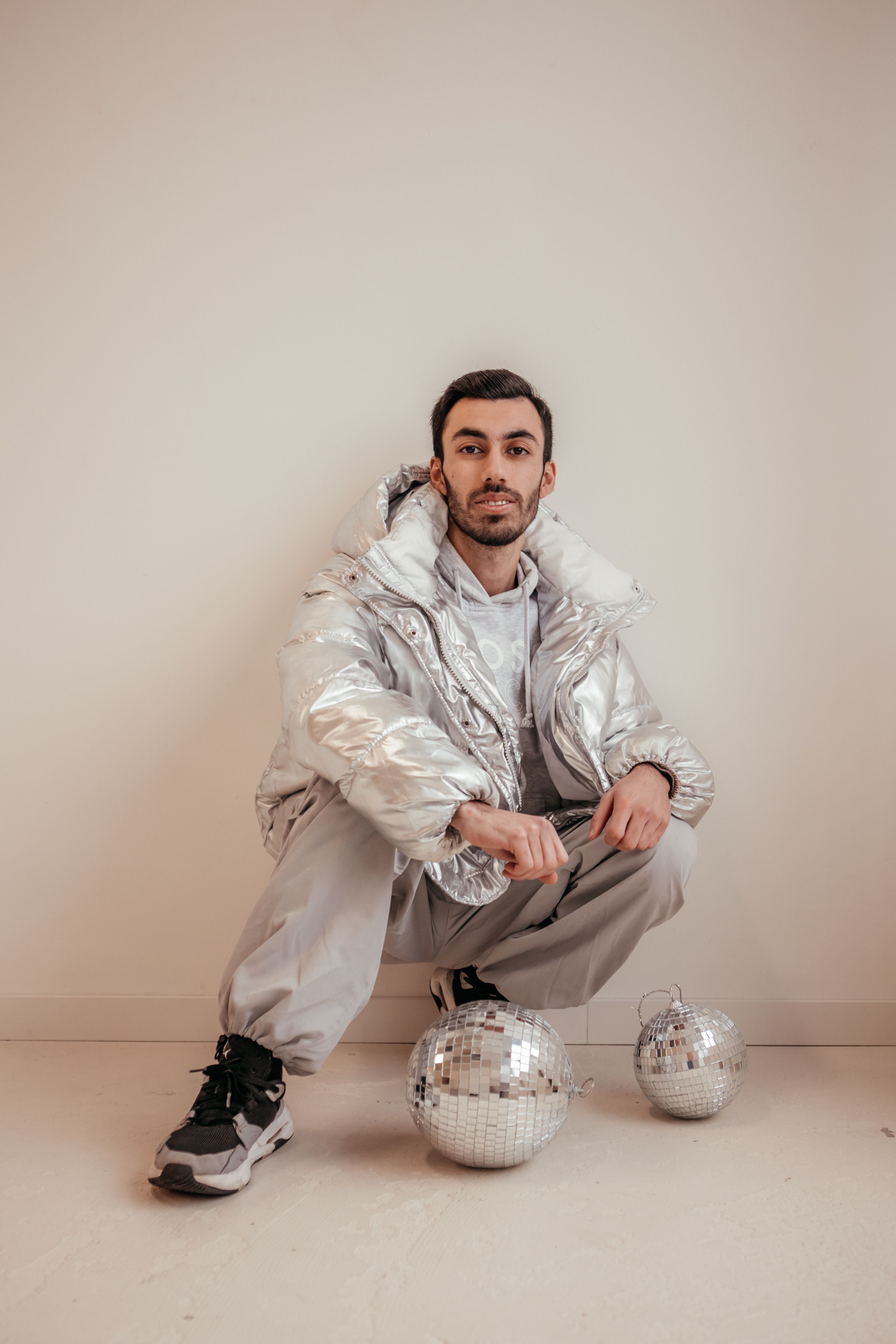 Stylish man wearing a silver jacket poses with disco balls indoors.