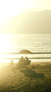 A family enjoying a sunny beach day under an umbrella, with a warm sunset over the ocean.