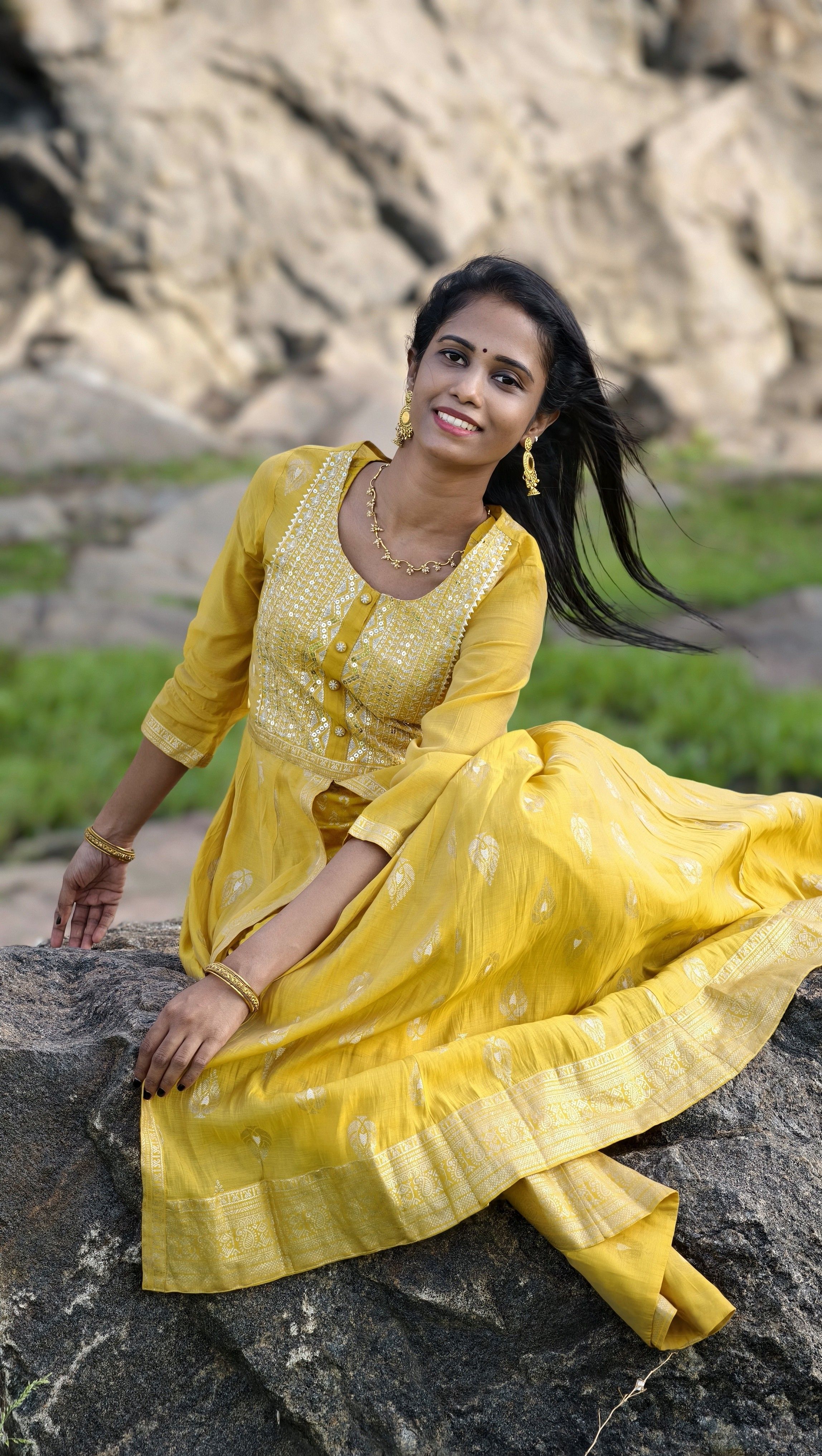 Smiling woman in vibrant yellow traditional dress sitting on rocks in nature.