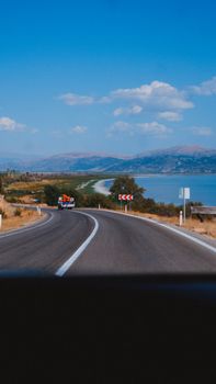 Curving road beside water, leading to distant mountains under a clear blue sky.