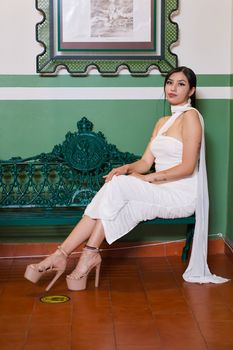 Fashionable woman in white dress seated on decorative green bench indoors, showcasing elegance.