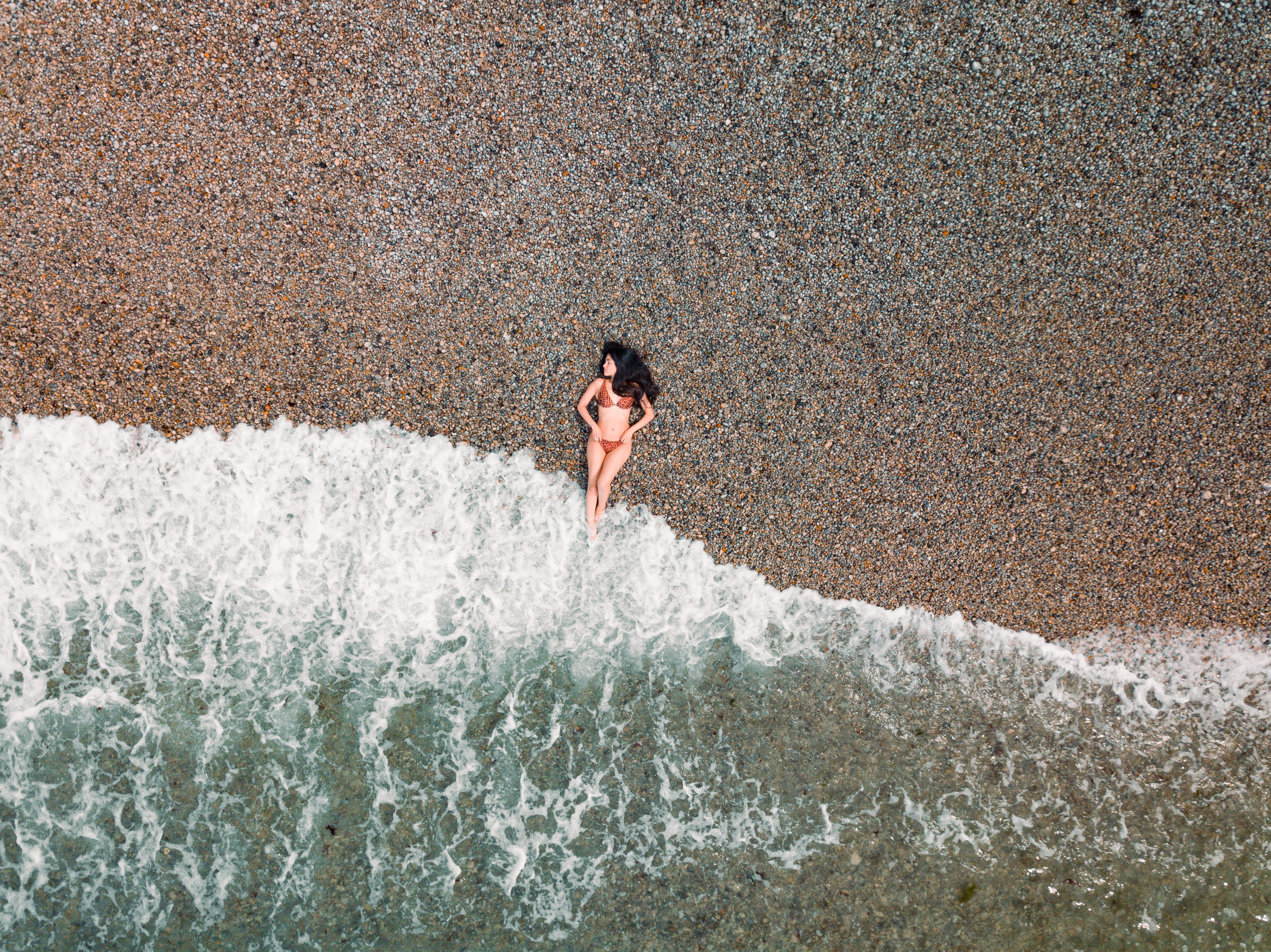 A top-down aerial shot of a woman in a swimsuit lying on a pebble beach with waves lapping at the shore.