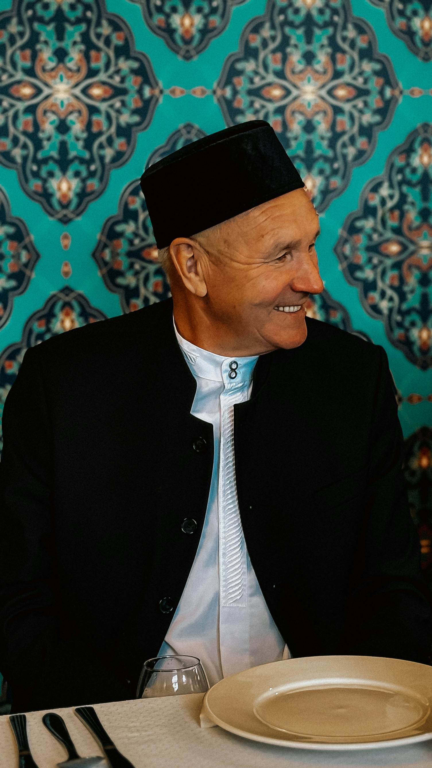 Elderly man wearing traditional black cap, smiling at dining table with decorative backdrop.