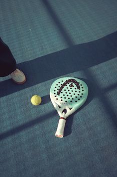 A padel racket and ball lie on a blue court in the sunlight, perfect for sports enthusiasts.