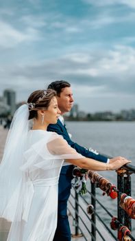 Bride and groom pose by a river, surrounded by cityscape and love locks, on a cloudy day.