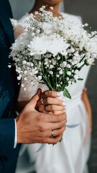 Close-up of couple's hands holding a white floral bouquet, highlighting wedding rings.