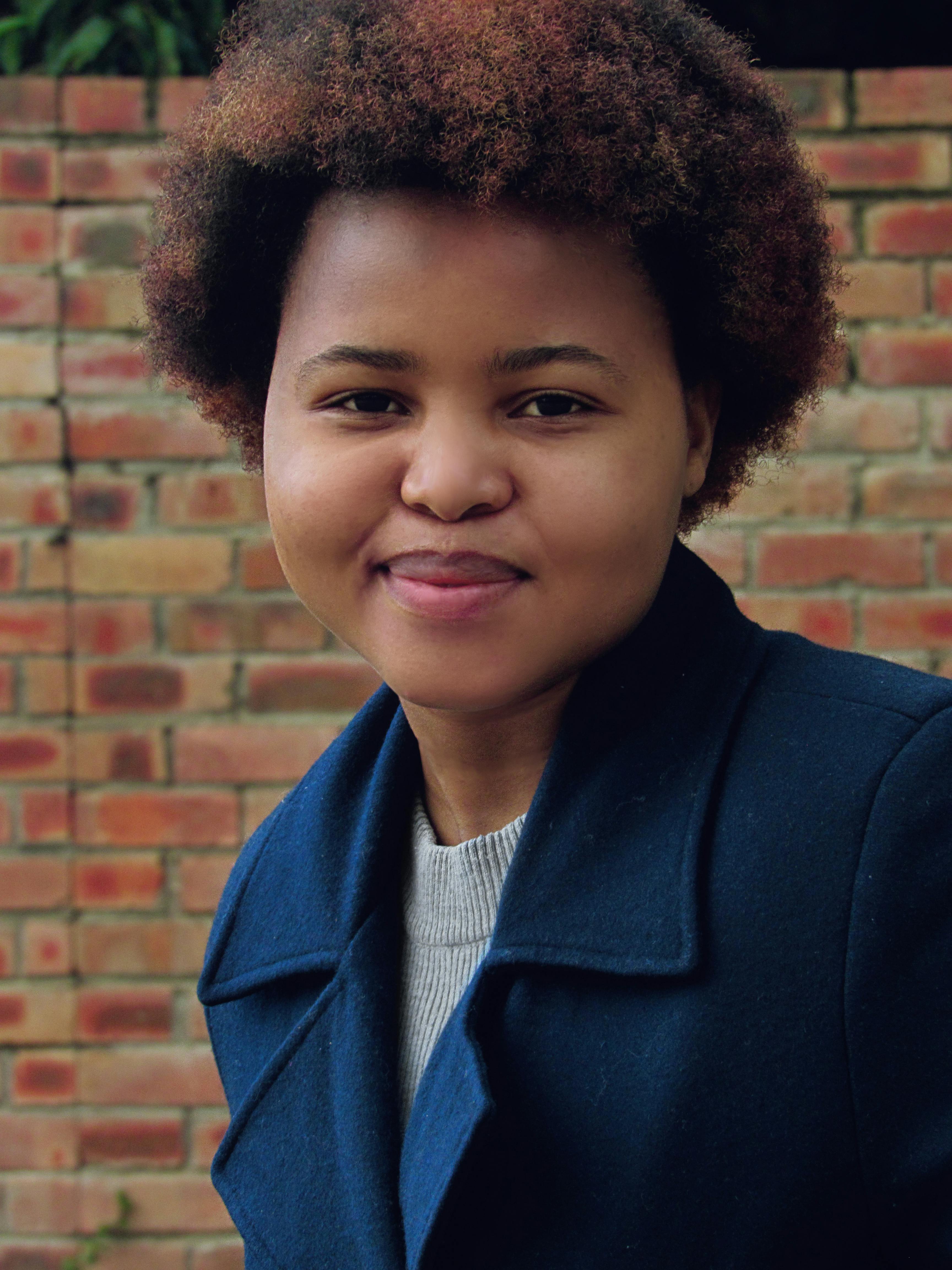 Portrait of a young woman in a navy coat smiling in front of a brick wall in Malkerns, Eswatini.