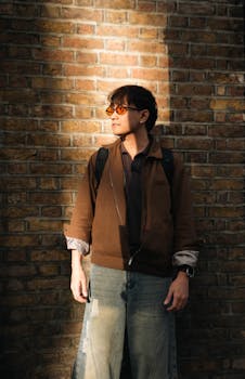 A fashionable young man in sunglasses stands against a brick wall in London. Afternoon sunlight creates a warm ambiance.