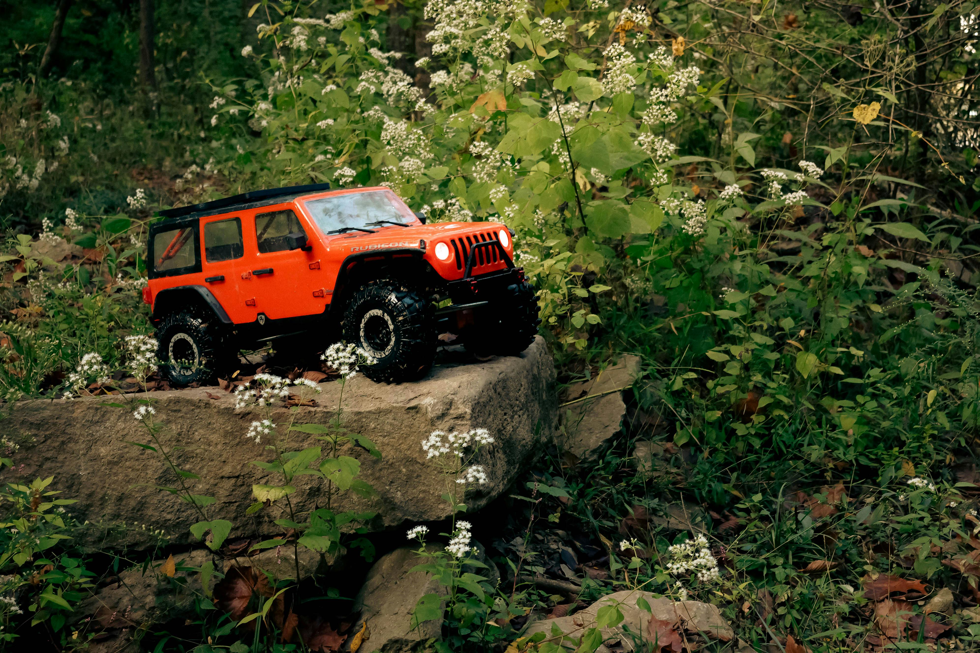 Orange toy Jeep on a rock in a lush forest scene, surrounded by greenery and flowers.