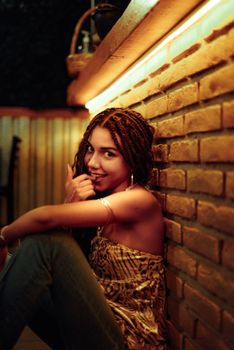 A young woman with braided hair sits against a brick wall in a warm, ambient indoor setting.