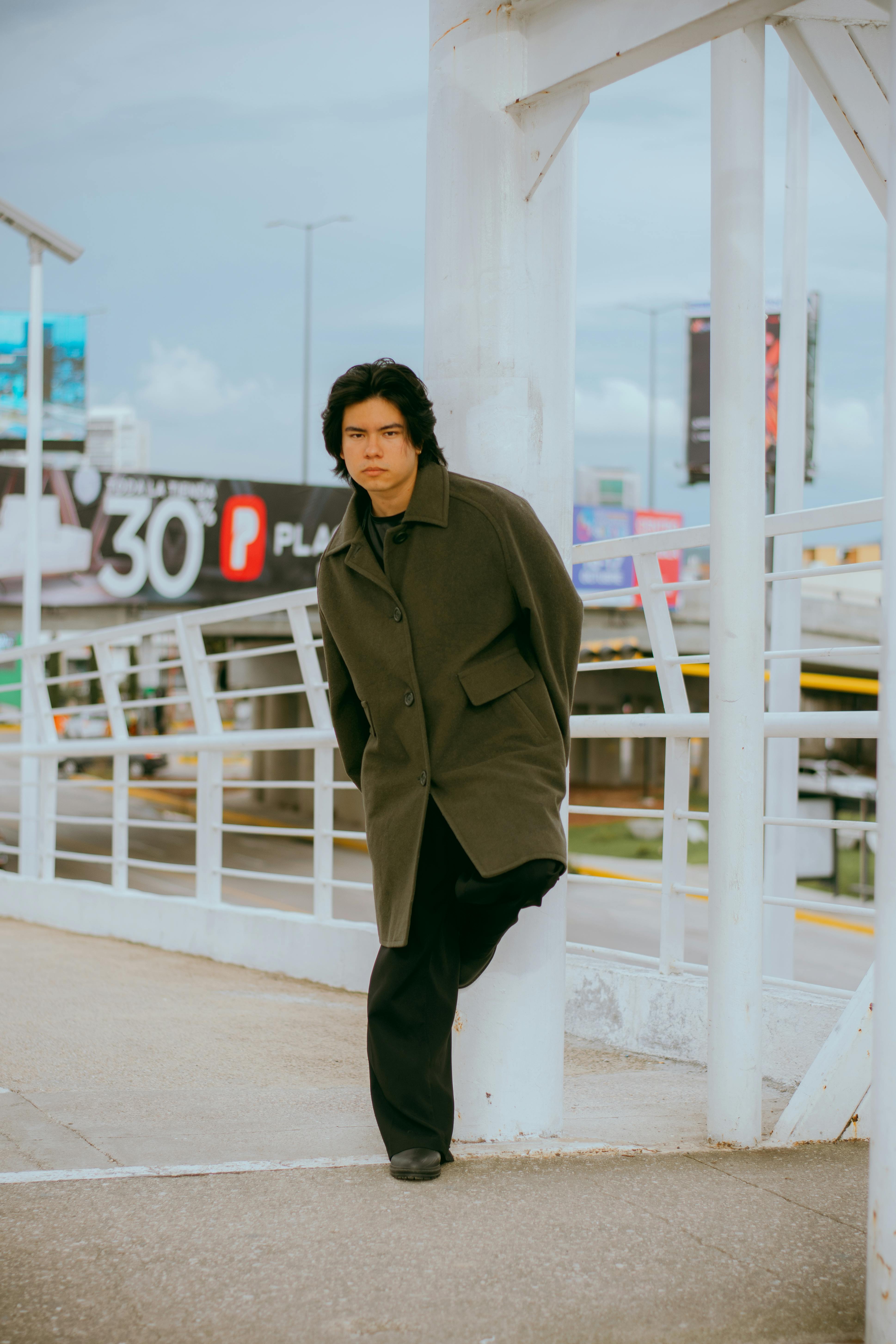 Young man in a coat leaning against a railing in an urban area.