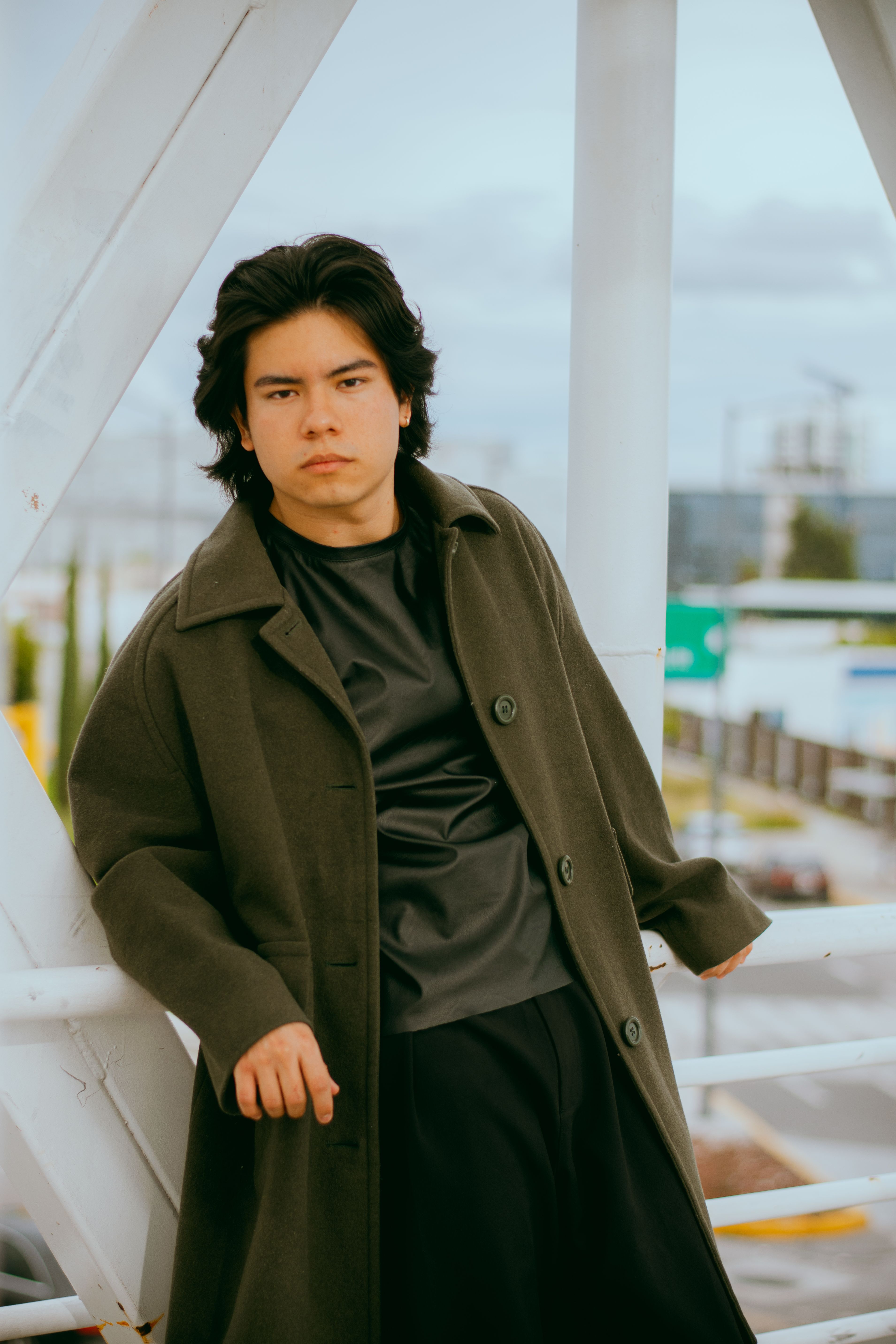 Stylish young man wearing a coat, leaning against an urban outdoor railing on a cloudy day.