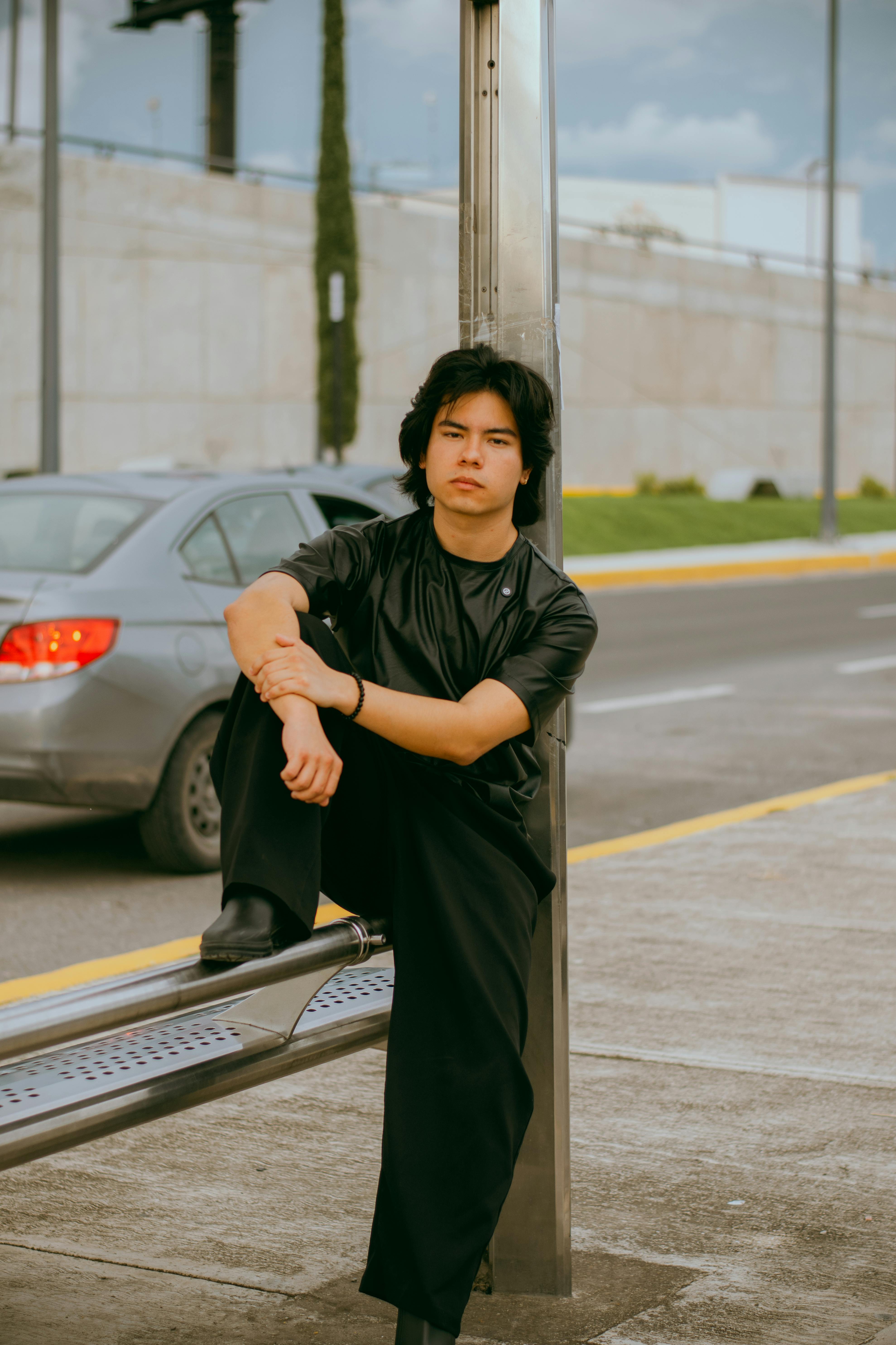 Young man sitting at a bus stop in a city setting, dressed in black. Urban lifestyle captured.