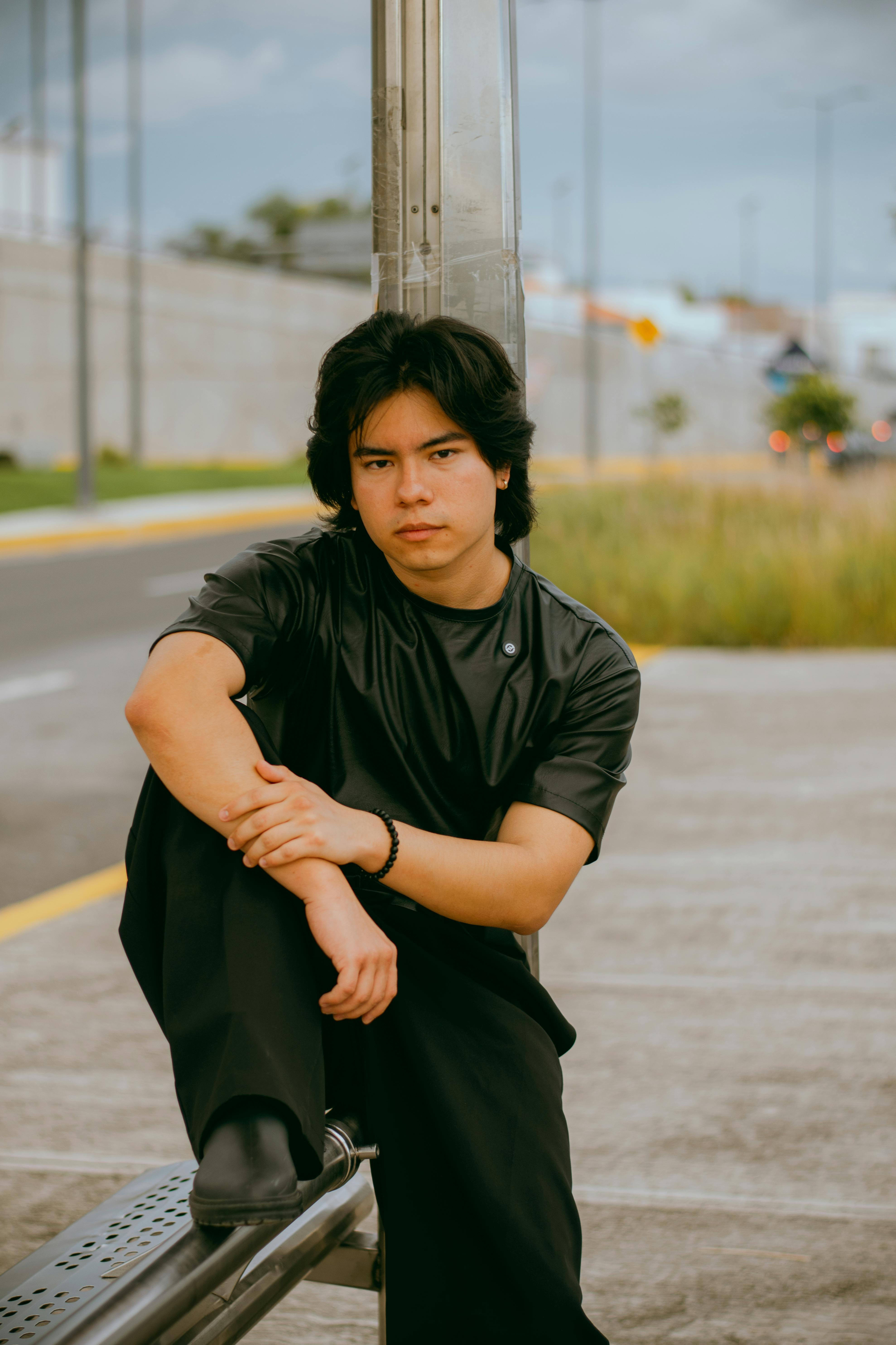 Contemplative young man in black attire seated outdoors on a sidewalk.