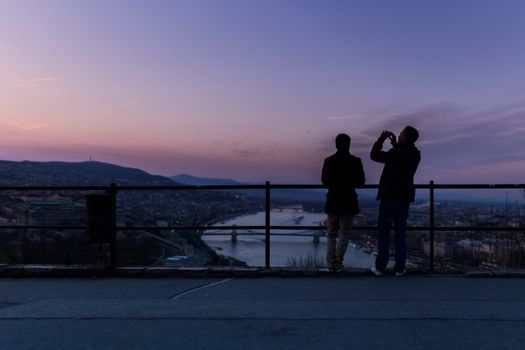 Silhouettes of two people capturing a stunning cityscape at sunset with a river view.