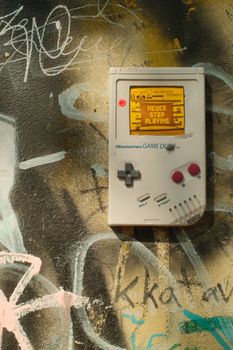 Retro handheld game console on graffiti-covered wall with 'Never Stop Playing' message displayed.