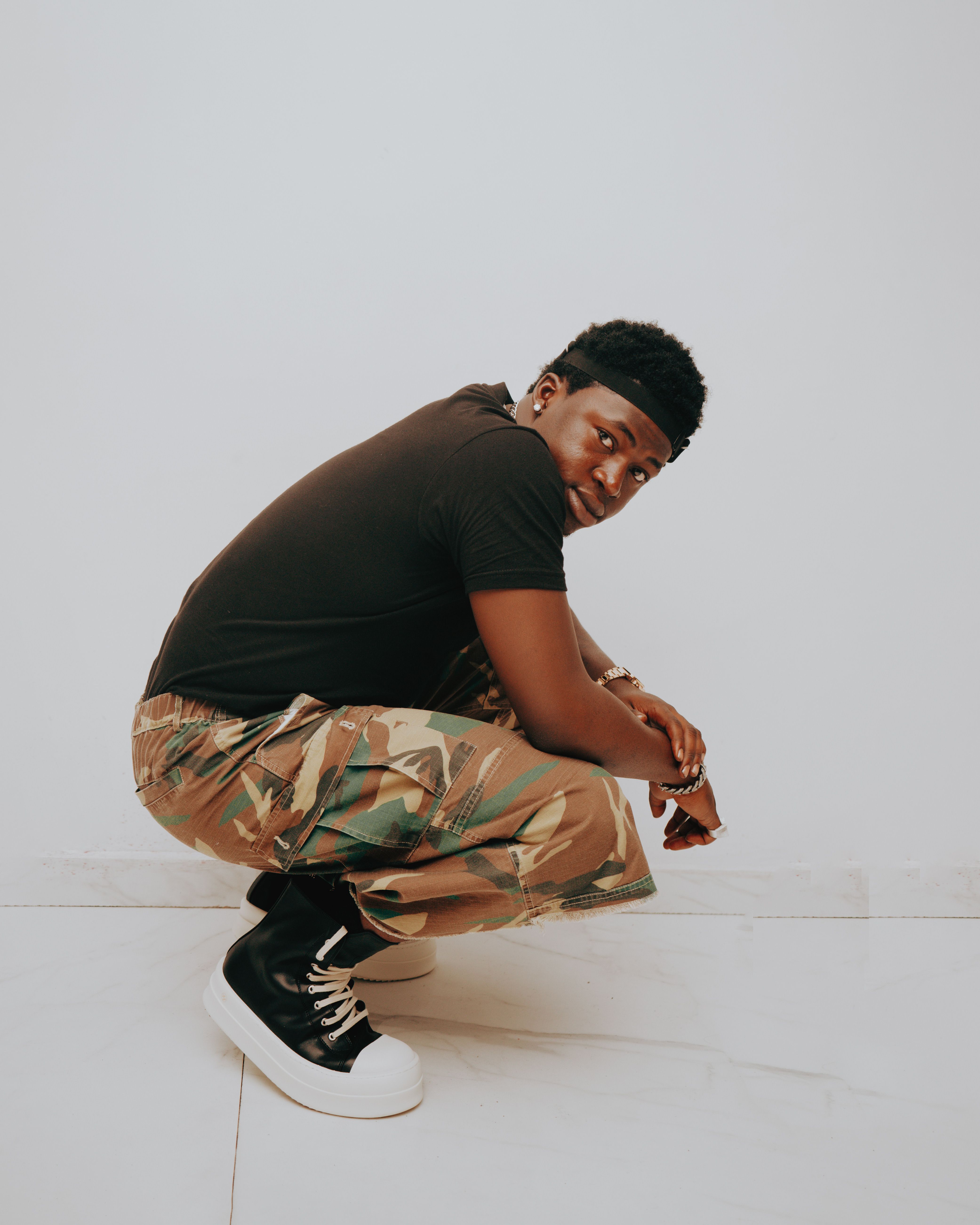 Stylish young adult wearing camouflage pants and black shirt in a crouching pose against a plain backdrop.