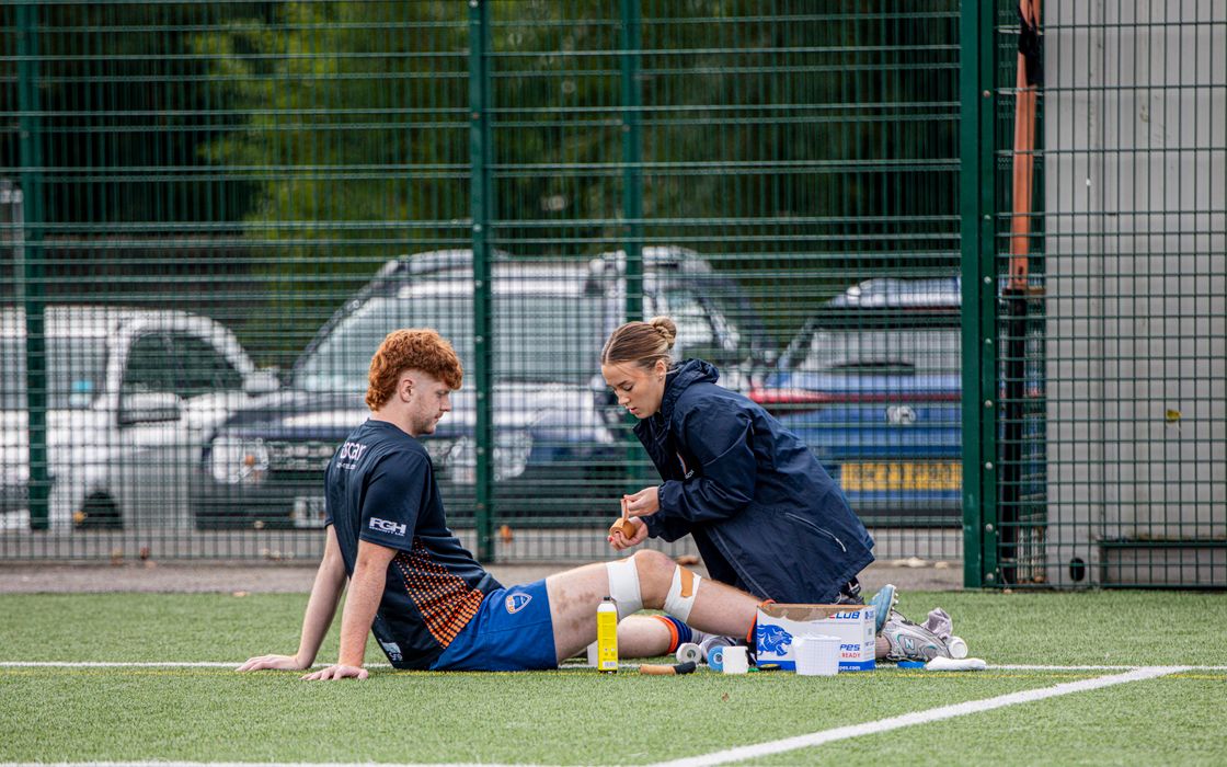 Sports therapist checking an injured football player sitting on an outdoor pitch