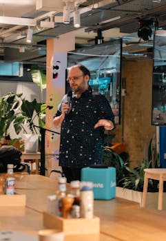 An adult male speaker with a microphone presenting indoors at a workshop event, surrounded by plants and modern decor.