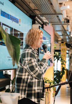 Blonde woman holding microphone and speaking at an indoor event with a presentation screen in the background.